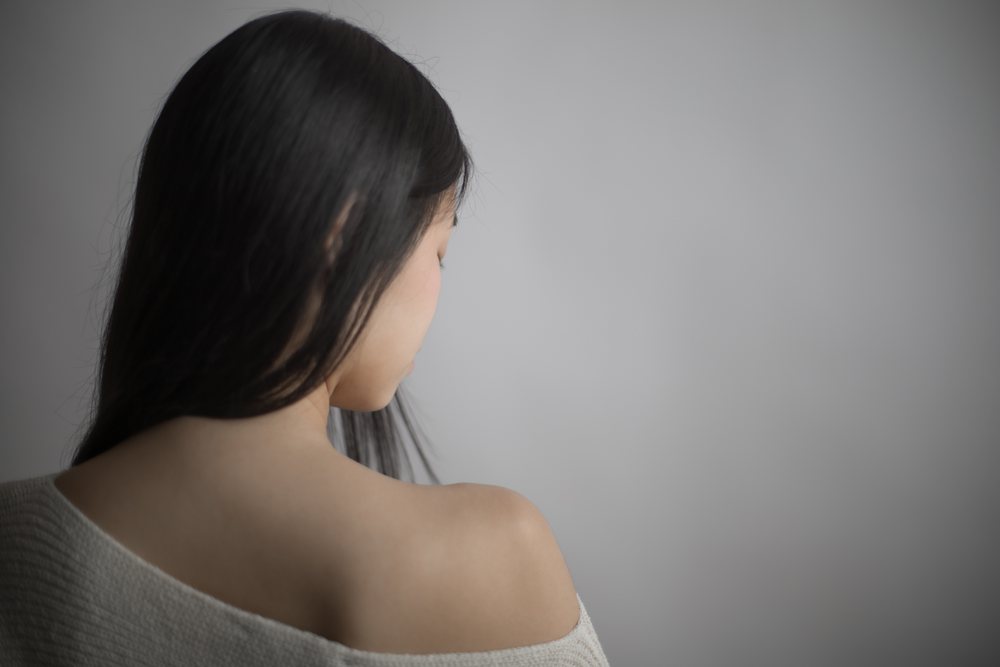 Woman with long black hair seen from behind, showing her back and shoulder in contemplation against a gray background, Art, portrait, emotional.