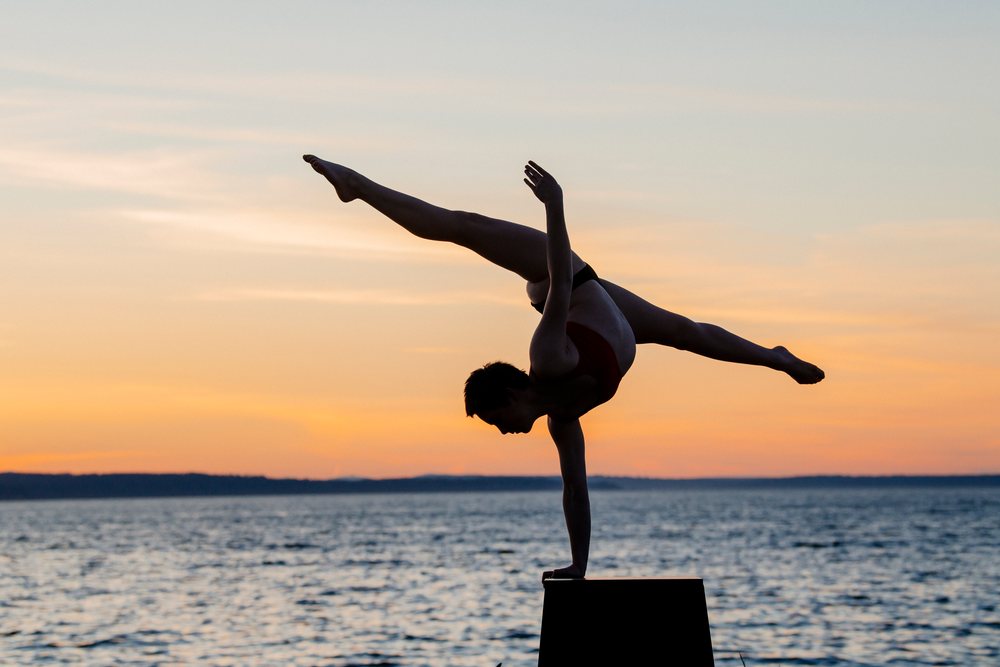 Female gymnast performing an outdoor handstand balancing on one hand by a lake at sunset, USA.