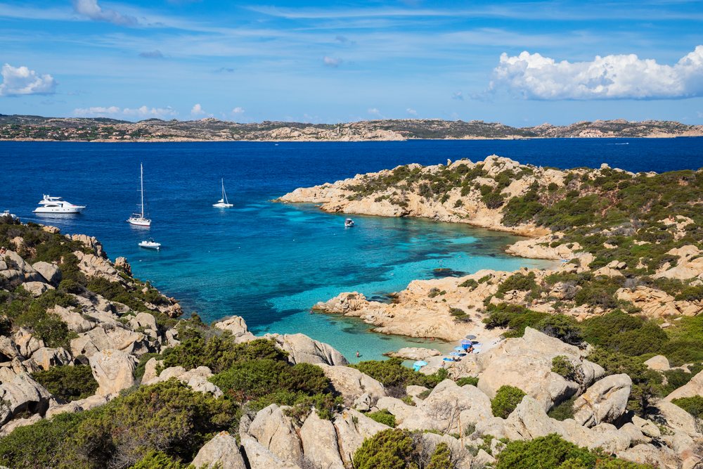 Cala Napoletana bay on Caprera Island features clear turquoise water, boats, rocky coastline, and Mediterranean coastal vegetation under a sunny sky with clouds, Travel Images, La Maddalena, Corsica, Italy.