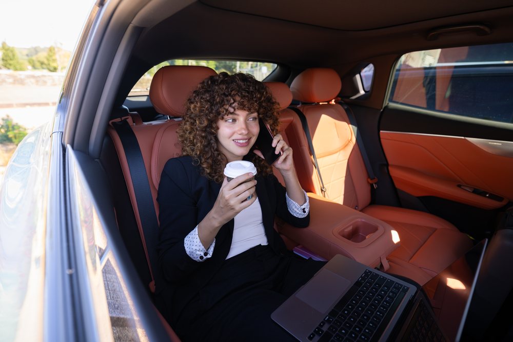 Businesswoman working remotely on a laptop and holding coffee while talking on phone inside a modern car in Spain during daylight, remote work, businesswoman, car.