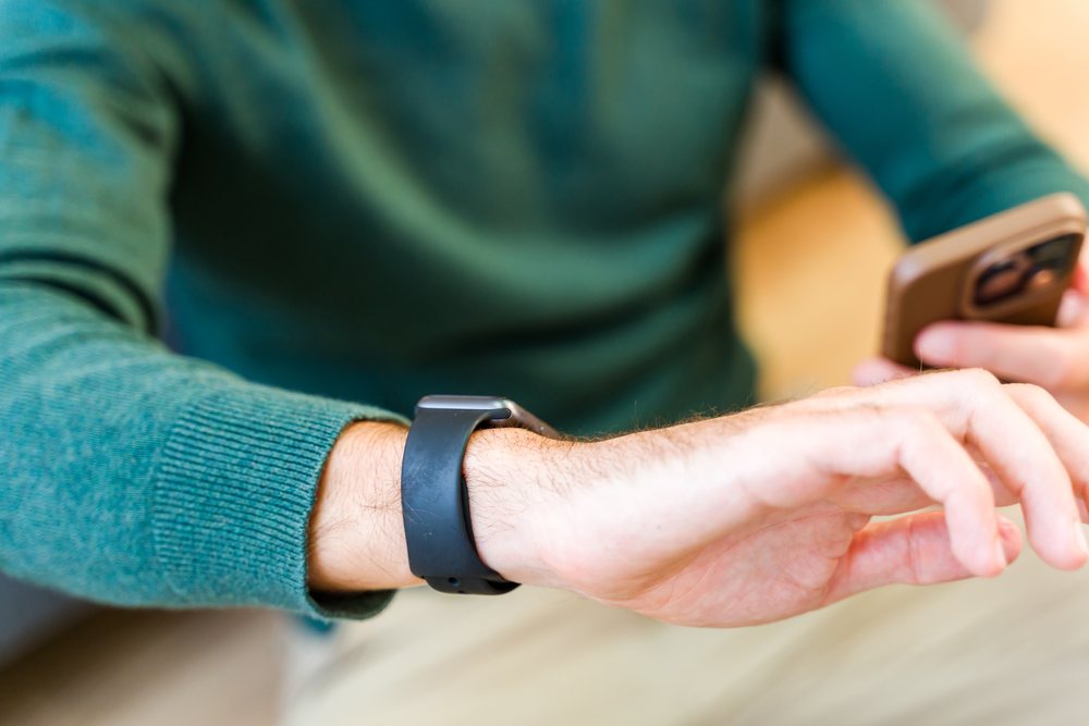 Man wearing a smartwatch and green sweater interacting with a smartphone indoors, Spain.