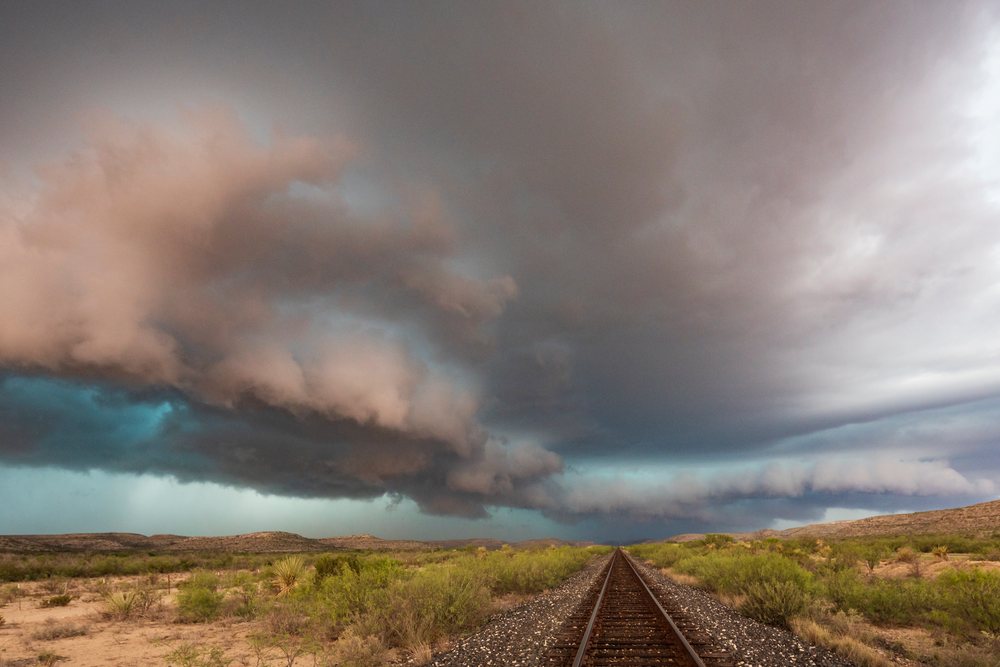 Railway tracks run through a flat, dry landscape of desert shrubs under heavy storm clouds in the Great Plains, Sanderson, Texas, USA.