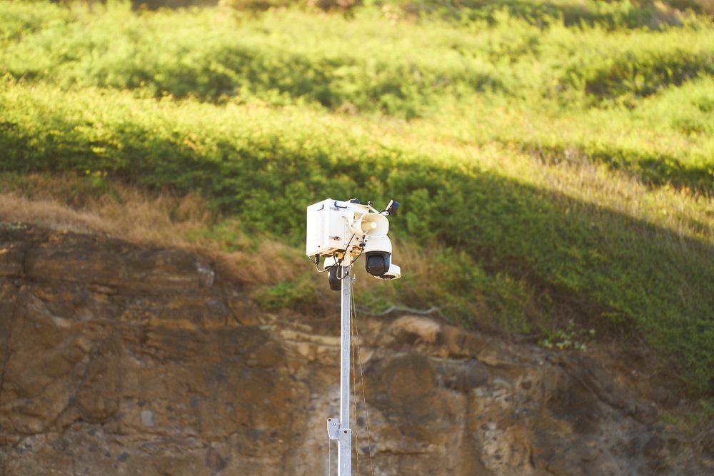 White security camera with multiple lenses mounted on a metal pole in front of a rocky hillside with green vegetation in daylight, Oahu, Hawaii, USA.