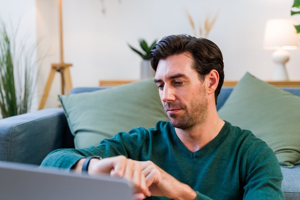 Man with brown hair wearing green sweater sitting on a couch looking at his wristwatch in a modern living room with pillows, lamp, and plants, Spain.