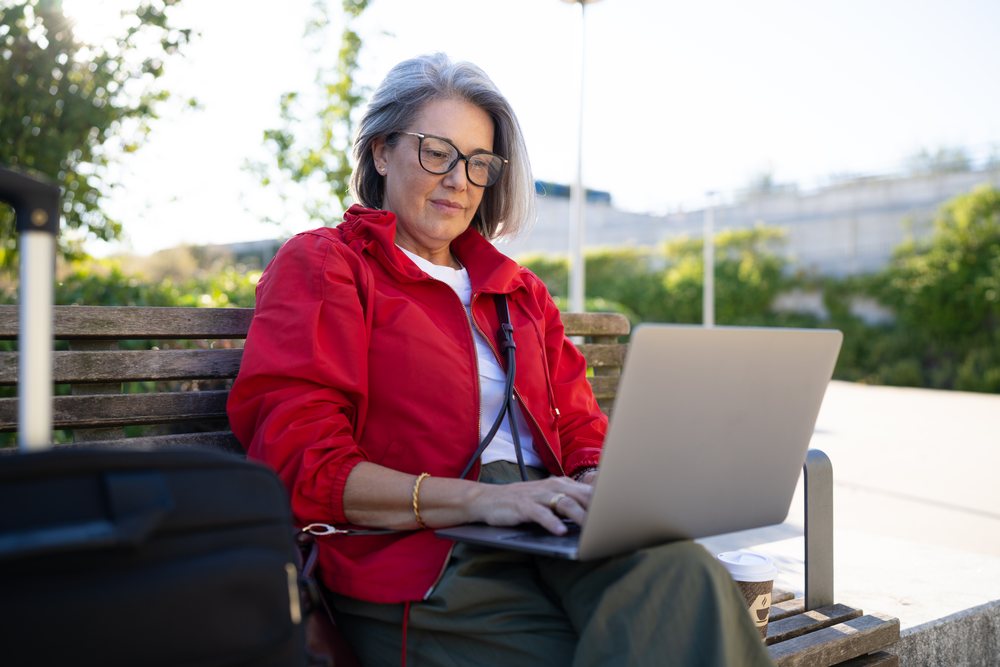 Woman wearing a red jacket working on a laptop while sitting on a wooden bench outdoors with a coffee cup and travel bag nearby, Spain.