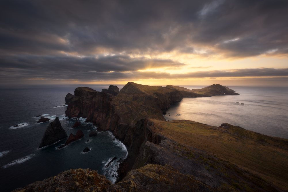 Rocky cliffs and ocean waters of Madeira Island illuminated by sunset sunlight under a dramatic cloudy sky.
