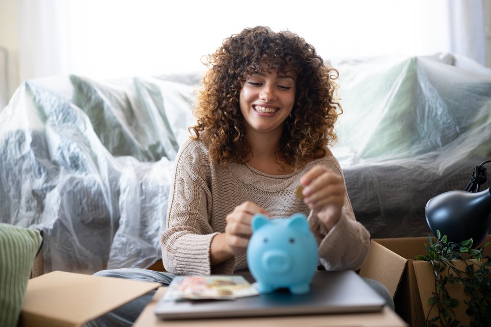 Woman smiles while inserting a coin into a blue piggy bank amid cardboard boxes and a plastic-covered couch during a move, moving, Spain.