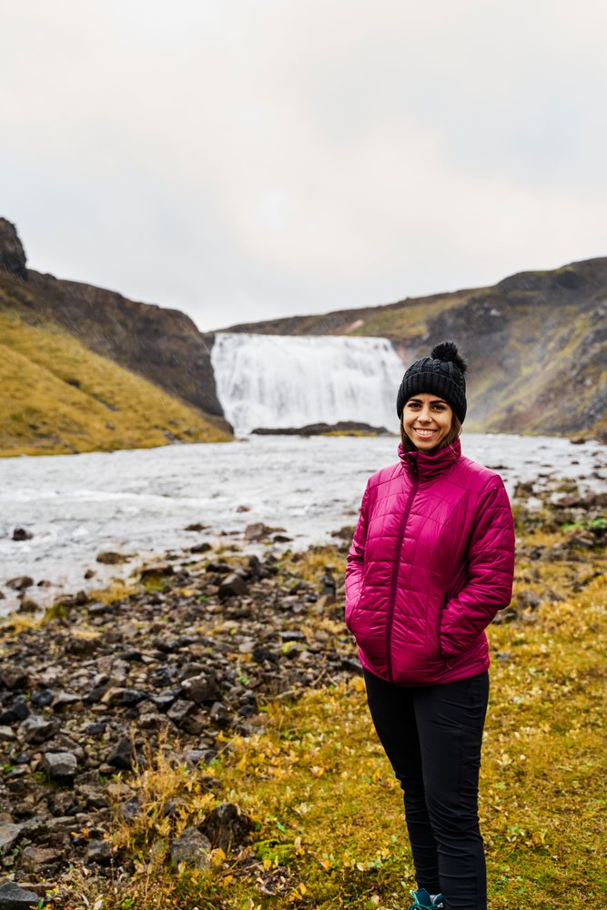 A woman wearing a magenta puffer jacket and black hat smiles while standing near a river with a waterfall in the background, Iceland.