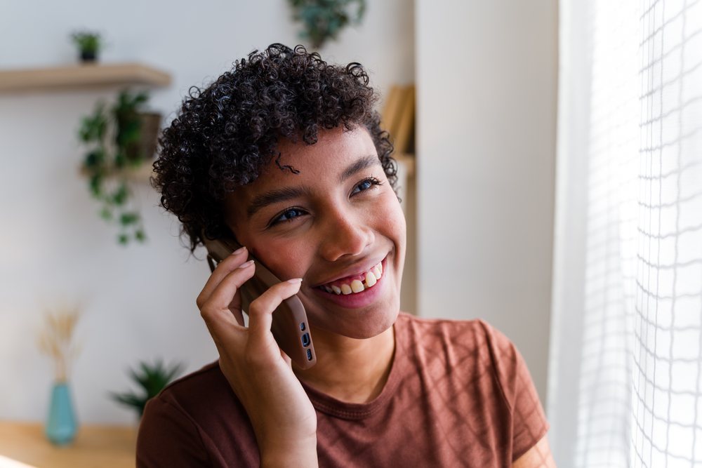 Woman with curly hair smiling while talking on a mobile phone indoors beside a window with natural light, Spain.