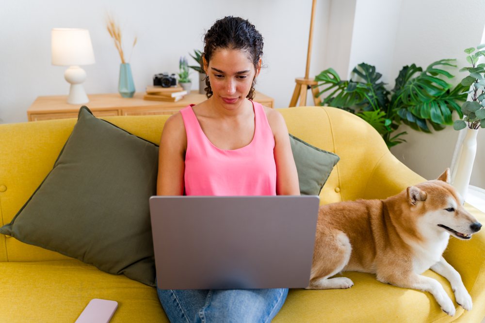 A woman working on a laptop while sitting on a yellow sofa with a small dog breed in a cozy indoor setting, Spain.