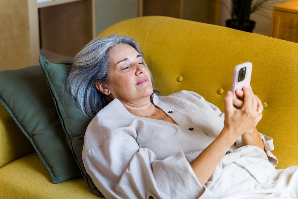 Middle-aged woman with gray hair relaxing on a yellow sofa holding a smartphone at home, Spain.