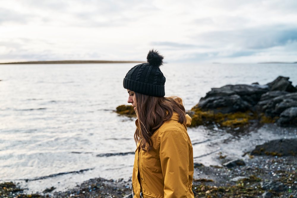 A white woman in a yellow jacket walks along a rocky shore