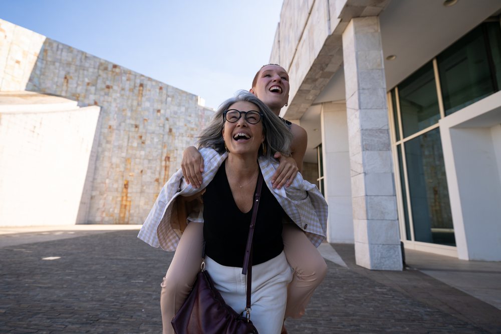 Mother gives her daughter a piggyback ride in an urban plaza outside a modern building, both laughing joyfully during the daytime, businesswomen, Spain.