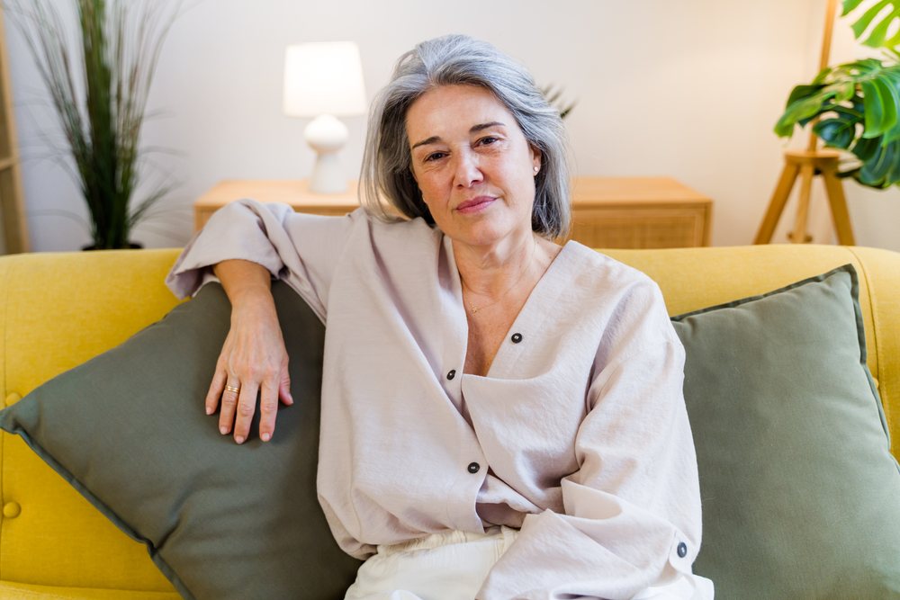 Middle-aged woman with gray hair sits relaxed on a yellow sofa with green cushions in a modern living room, Spain.