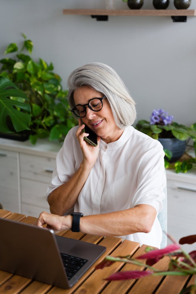 Mature woman checking the time on a wrist device while talking on the phone and working on a laptop indoors, Spain.