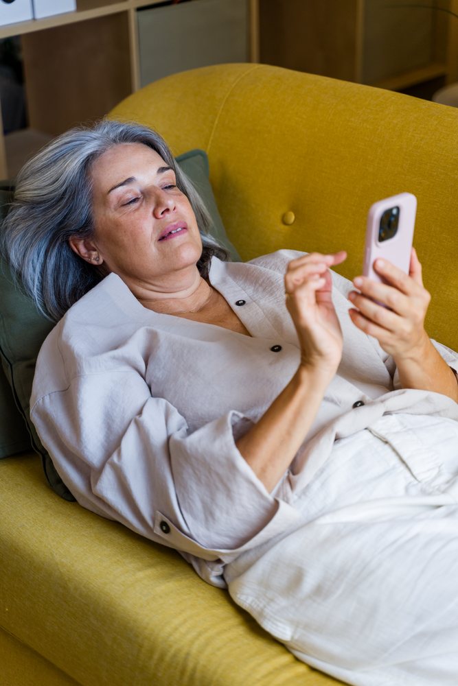 Woman with gray hair lying on a yellow couch indoors using a smartphone, Spain.