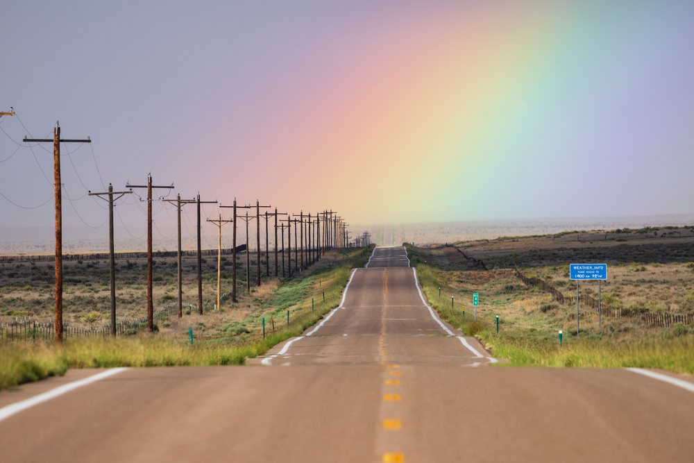 Rainbow arching over Highway 10, with telephone poles and a blue road sign alongside the asphalt road, Storm Chasing, Weather, Environment, Rocky Ford, Colorado, USA.