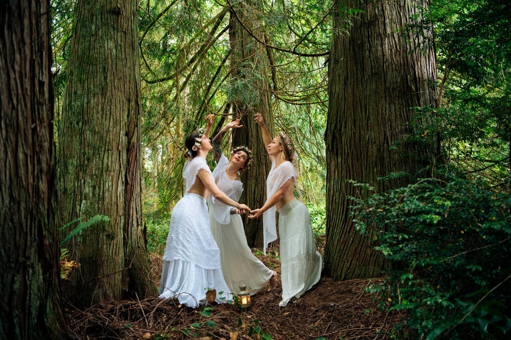 Three women in white dresses wearing flower crowns dance together in a green forest, muses, USA.