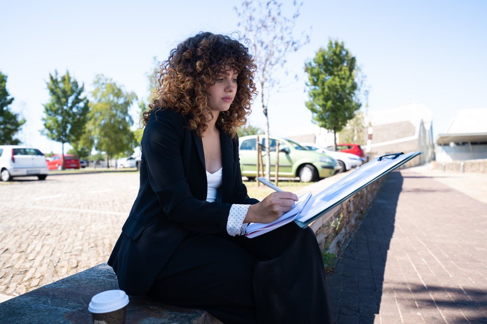 Businesswoman sitting outdoors in a parking lot in Spain writing in a notebook with a clipboard and coffee cup nearby, businesswoman, traveling executive.
