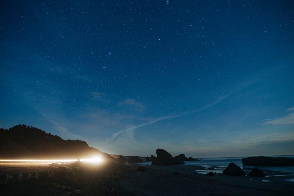 Stars fill the night sky above coastal rock formations, ocean waters, and a curving highway lit by car headlights, Meyers Creek, Oregon, USA.