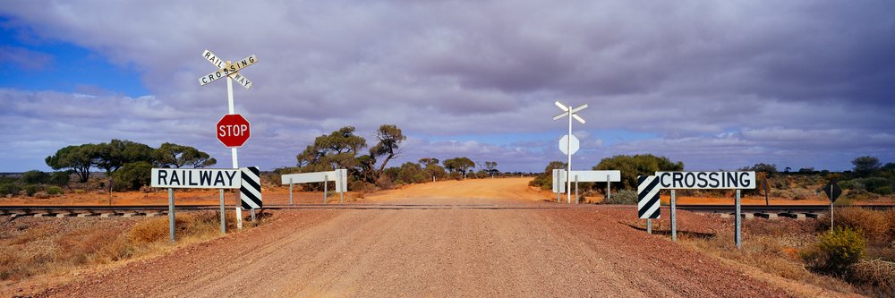 Railway crossing with stop and crossbuck signs on a dirt road in the arid, bushland landscape of Outback Australia under a cloudy sky.