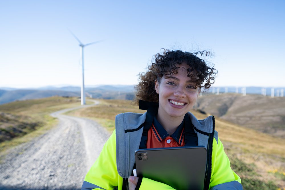 Adult woman in a hi-vis jacket holding a tablet stands outdoors on rural hills with multiple wind turbines visible in the background, sustainability, renewable energy, Galicia, Spain.