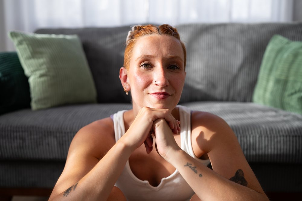 A young woman with red hair and tattoos sits indoors on the floor resting her chin on her clasped hands in a living room with a gray sofa and green cushions, moving, Spain.