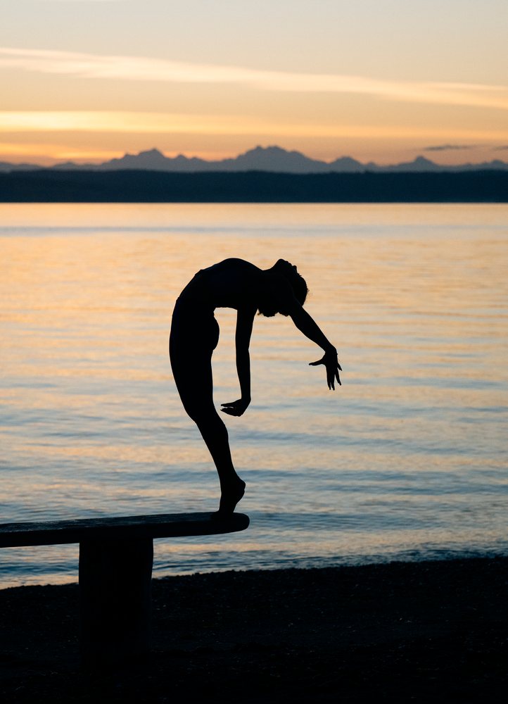 Woman practicing yoga acrobatics in silhouette on a bench by the water at sunset, USA.