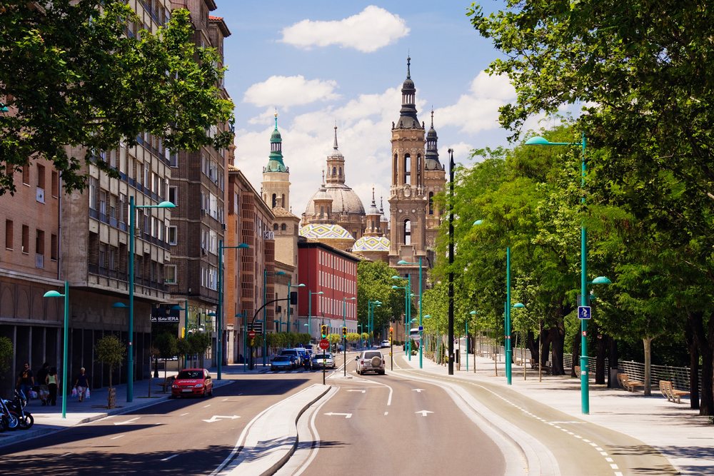 Street view in Zaragoza shows a cathedral with towers and domes, cars and pedestrians on a tree-lined road under a blue sky with clouds, Aragon, Spain.