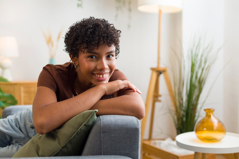 Young woman with curly hair smiling while resting on a grey couch.