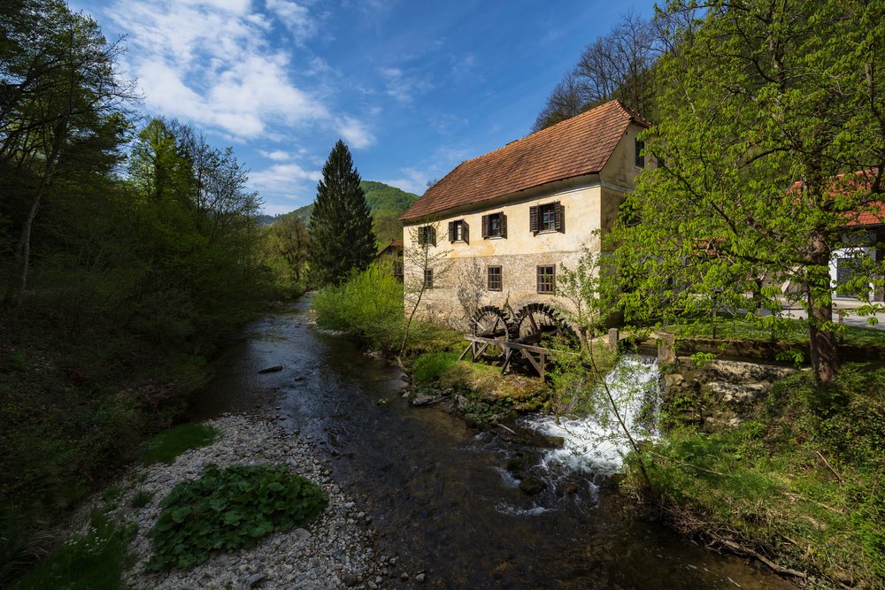 Mill with wooden water wheel beside a flowing stream in a lush green forest, landscape, cityscape, Podsreda, Slovenia.