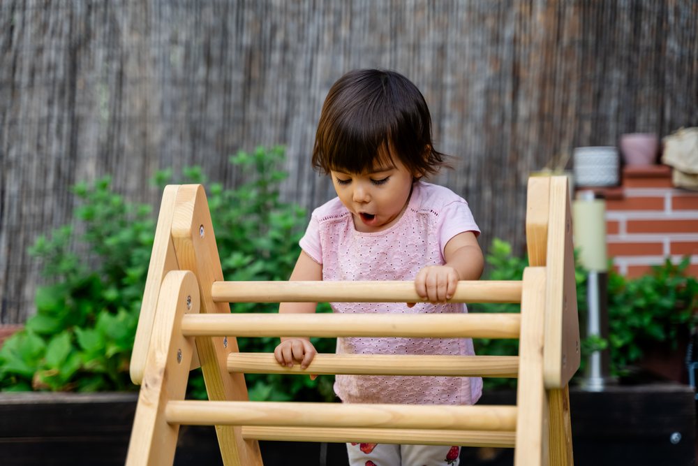 Toddler girl playing on a wooden climbing frame in a garden outdoors showing curiosity and wonder, kids, Spain.