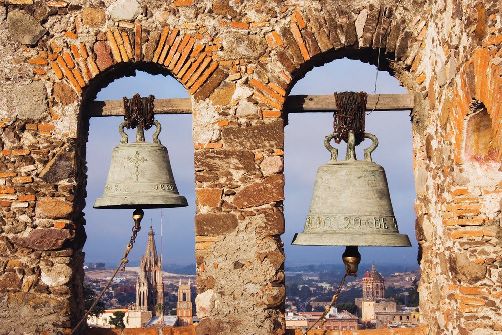 Church bells hang in a stone belfry with San Miguel de Allende city buildings and skyline visible in the background, Guanajuato, Mexico.