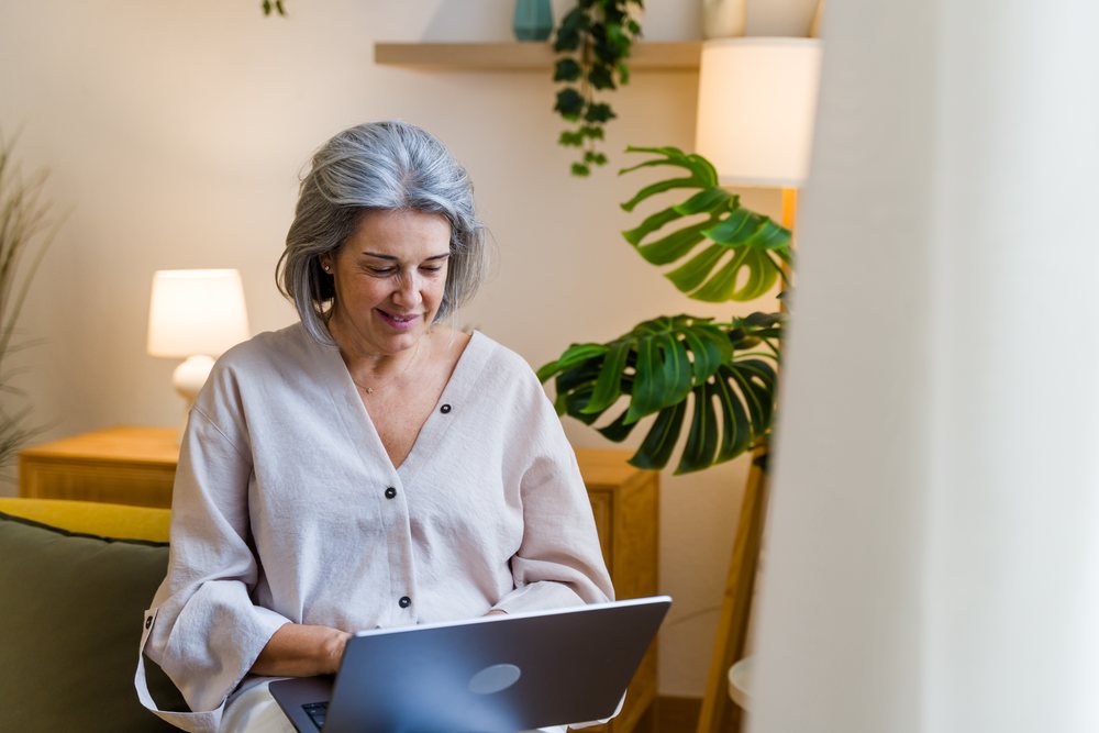 Mature woman with gray hair smiles while sitting indoors using a laptop near plants and lamps, Spain.