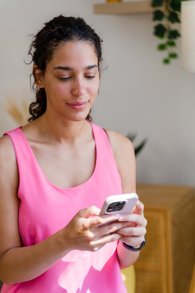 A young woman in a pink tank top uses her smartphone indoors in a home setting, Spain.