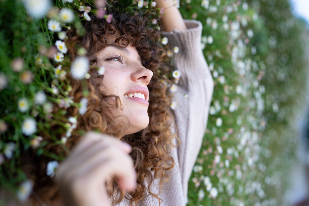 Woman with curly hair lies smiling in a natural daisy field outdoors in Spain under daylight, home story, influencer.