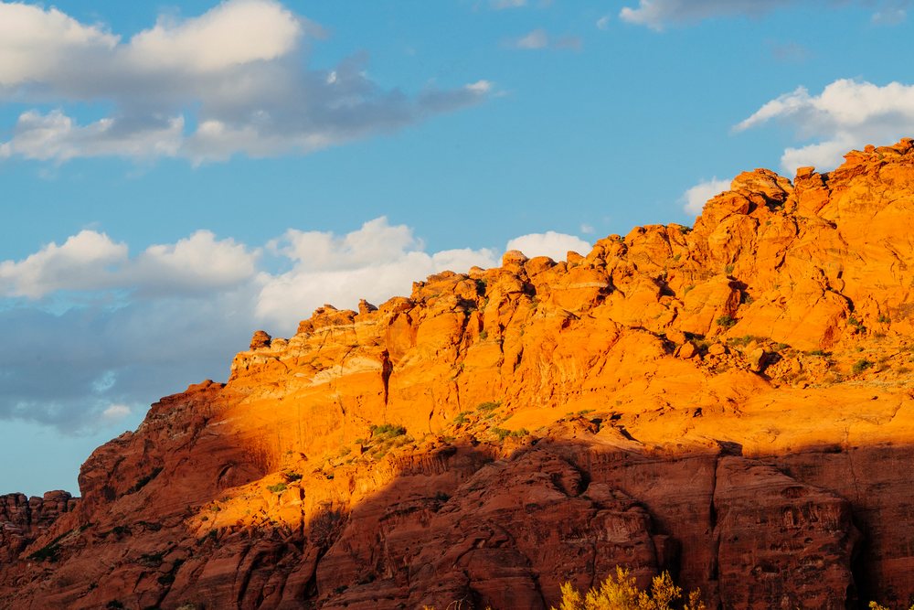 Sunlit red cliffs with yellow foliage in red Cliffs Desert State Park, under a partly cloudy blue sky, Snow Canyon, Utah, USA.