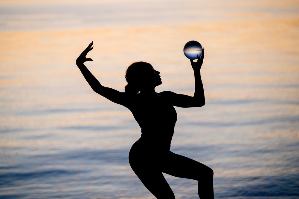Silhouette of a woman balancing a clear sphere while performing a yoga acrobatic pose at sunset by the water, USA.