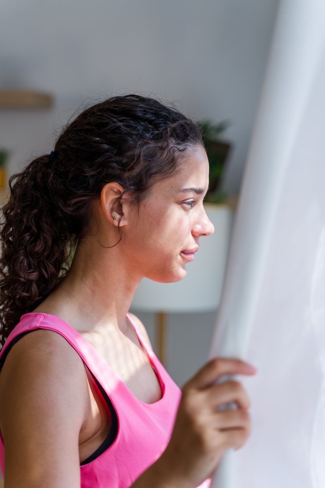 A young woman in a pink tank top looks thoughtfully out the window while holding a white curtain inside a home, Spain.