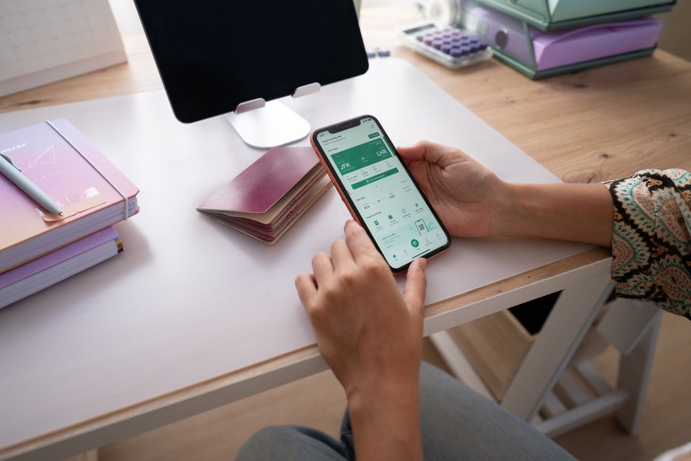 Person holding a smartphone showing a digital flight boarding pass beside a passport, notebooks, and pen on a wooden desk in a home office, Spain.