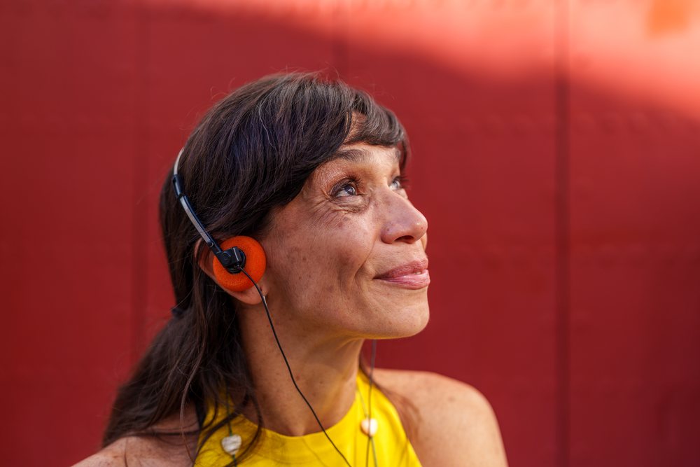 Mature woman wearing orange on-ear headphones and a sleeveless yellow top smiles while listening to music outdoors against a red wall, retro, vintage, Málaga, Spain.