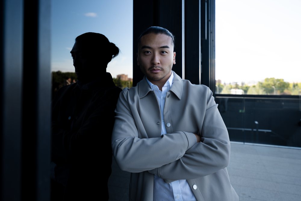 Man standing outdoors with arms crossed and reflection in a glass window of a modern building, Madrid, Spain.