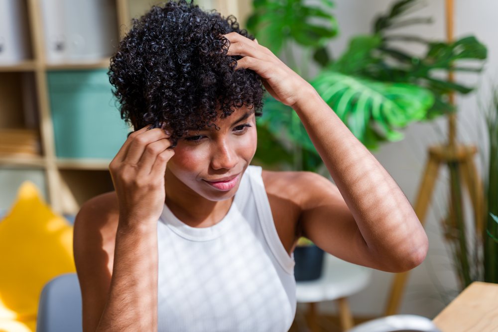 Young woman indoors adjusts her curly black hair with green plants in the background, Spain.