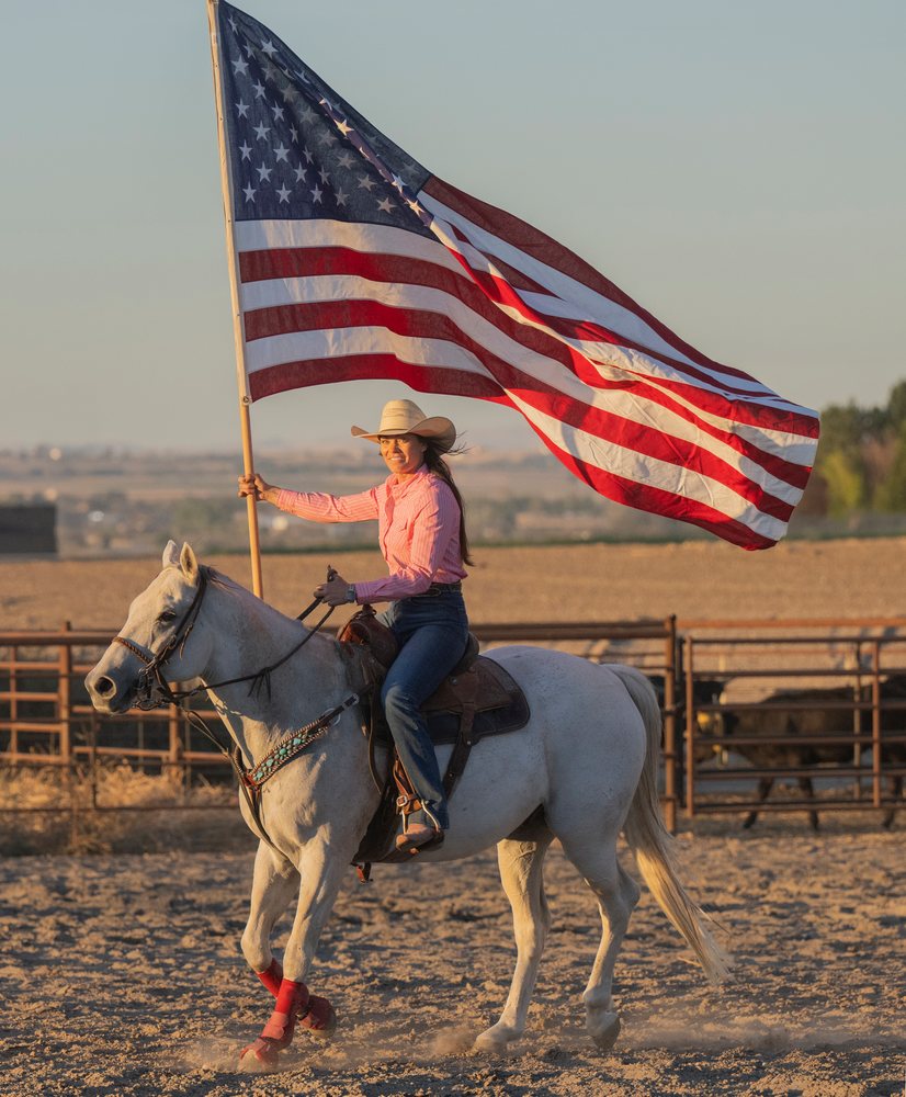 Woman wearing a cowboy hat rides a white horse with red front wraps, holding a large American flag in a dusty rodeo arena at sunset, Southwest USA.