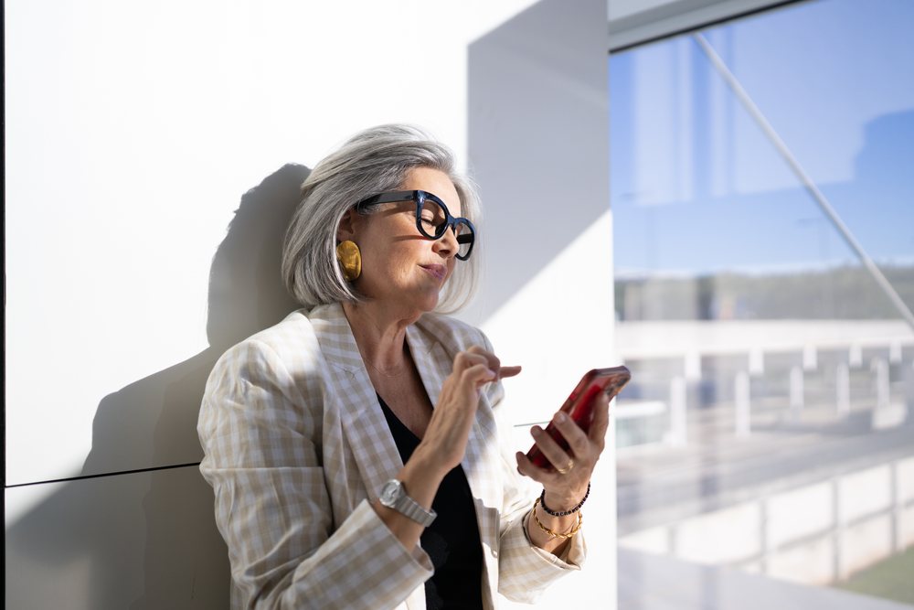 Businesswoman with gray hair using a smartphone while leaning against a wall inside a modern building, Spain, Madrid.
