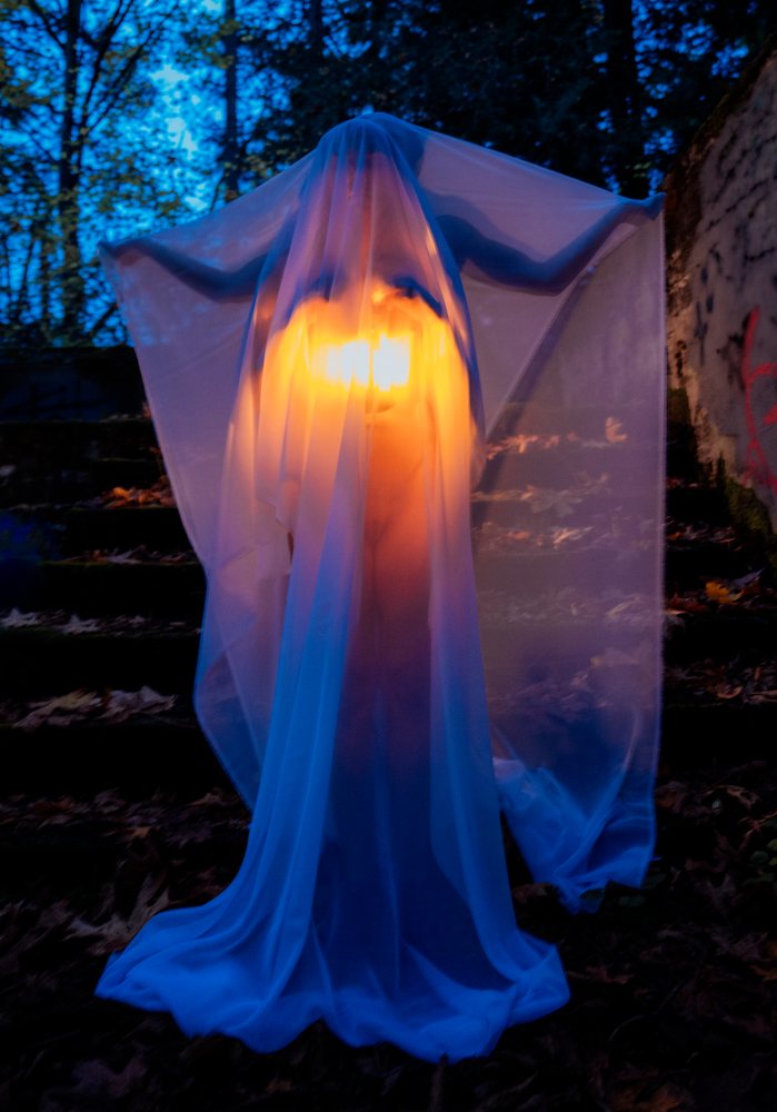 Ghost figure draped in white translucent fabric glows with orange light outdoors at night among trees and fallen autumn leaves, ghosts, USA.