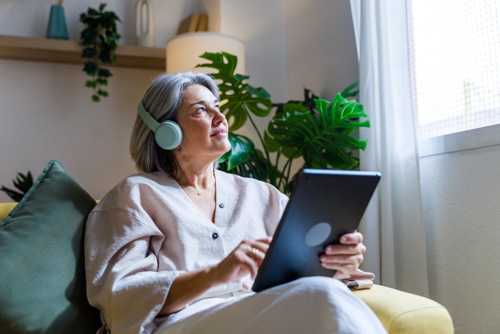 Gray-haired woman wearing headphones sits on a sofa using a tablet while looking out a window in a cozy room with indoor plants and natural light, Spain.