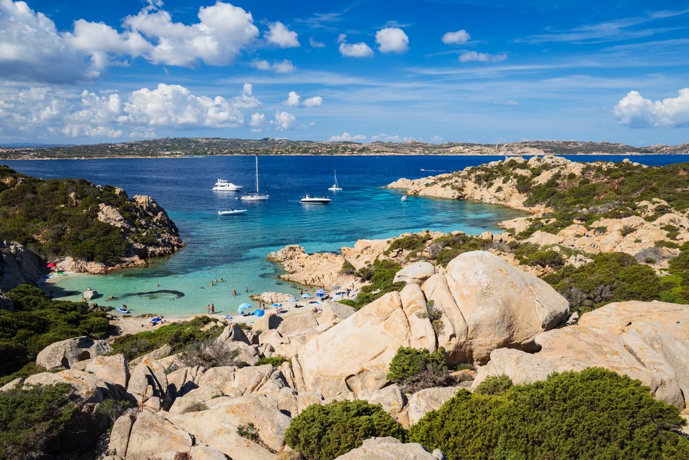 People swim and sailboats anchor in a capsized cove at Cala Napoletana, coast of Caprera Island, La Maddalena, Sardinia, Italy.