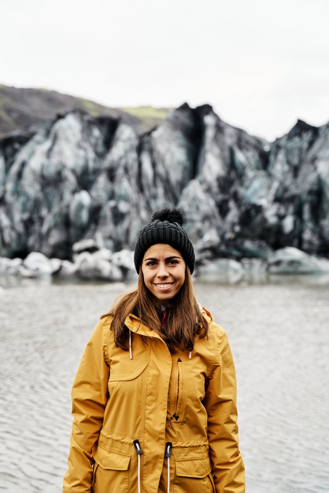A white woman in warm clothes standing by a glacier lake