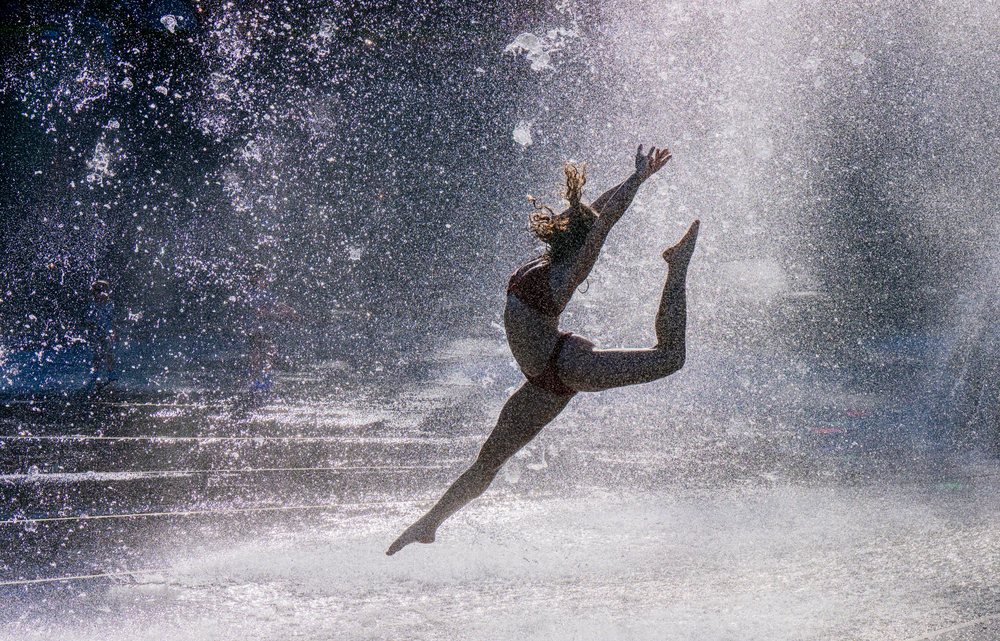 Woman jumping energetically through water sprays from an outdoor fountain in the USA on a sunny day, partner, acrobatics, yoga.